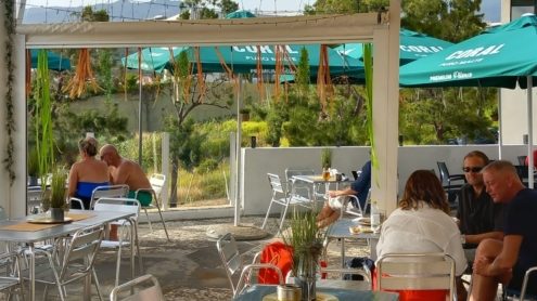 Metal tables and chairs on the covered terrace at Charlotte's Place in Funchal, green parasols over them, people sitting out and a wooded slope beyond.
