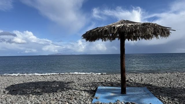 A thatched straw parasol on a blue concrete base standing on an empty pebble beach, with the Atlantic and low cloud beyond