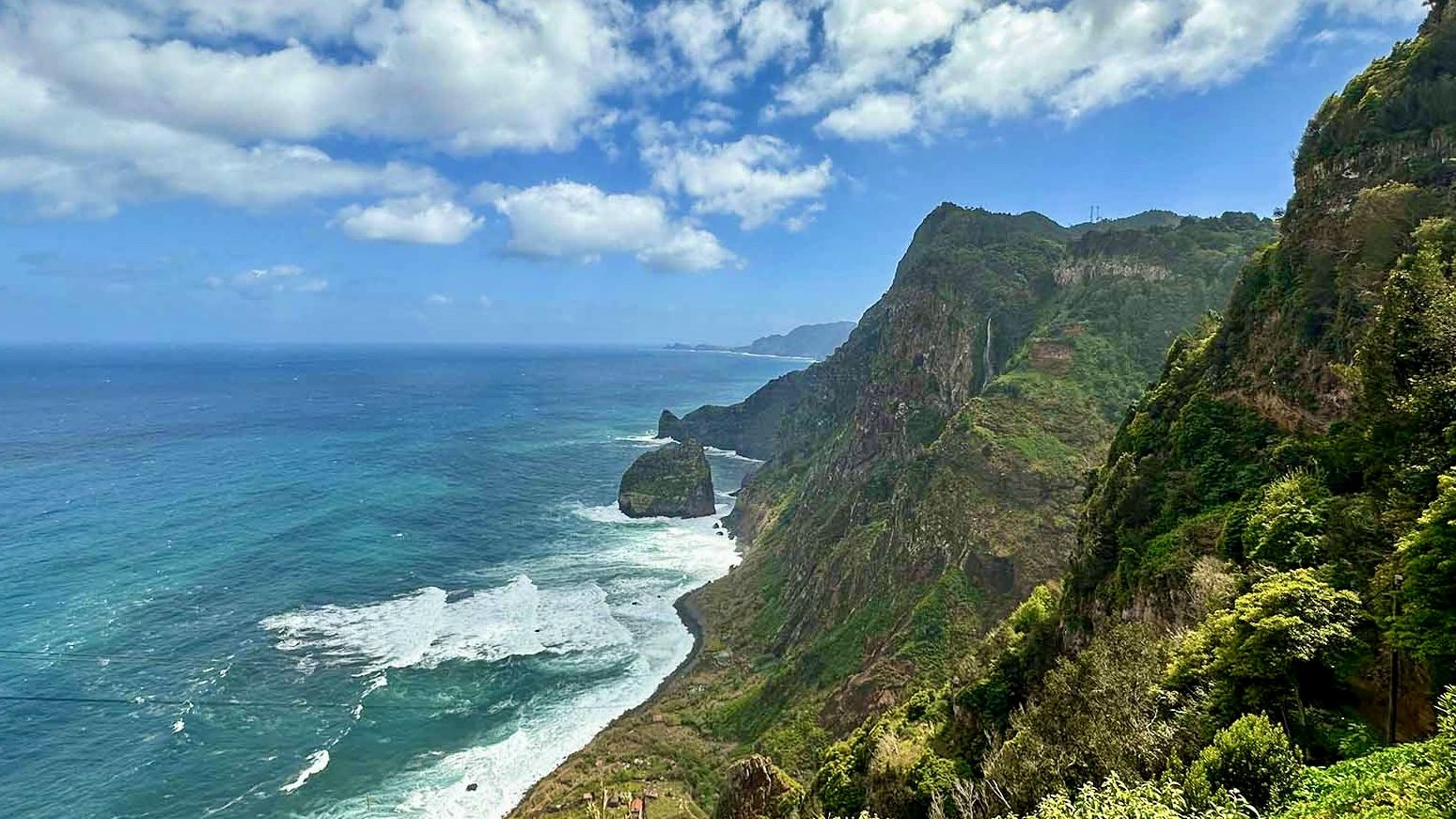Green Madeira cliffs falling to the Atlantic, with a tall sea stack standing in the white surf and a thin waterfall on the rock face