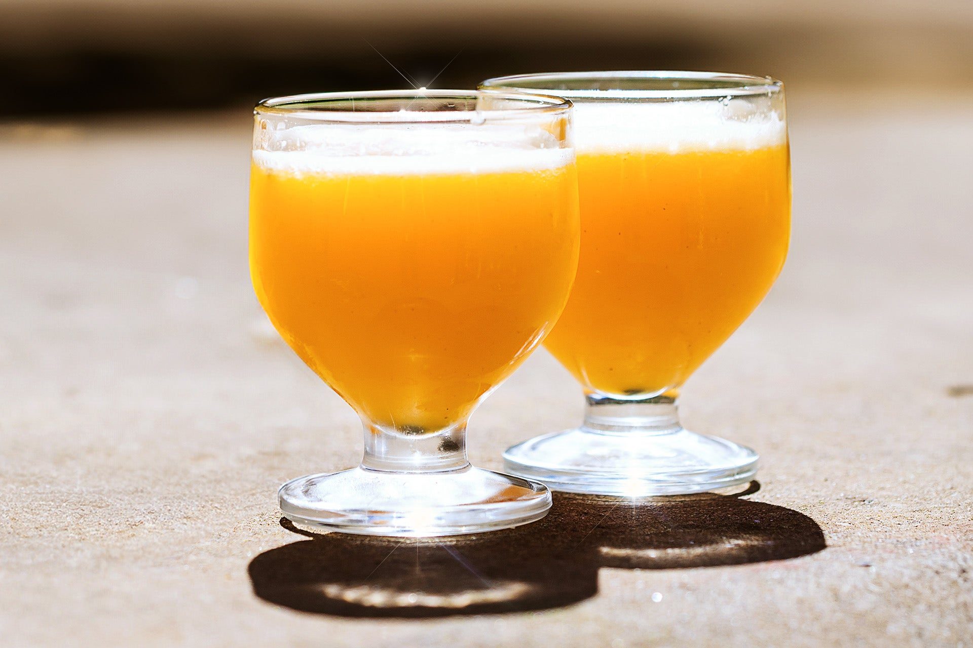 Two small stemmed glasses of golden poncha with a froth on top, standing in sunlight on a stone surface