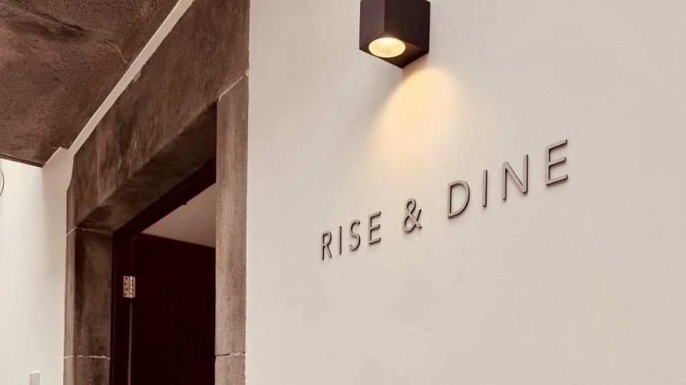 Raised lettering spelling Rise & Dine on a white wall beside the basalt doorway of the brunch room on Rua da Carreira in Funchal