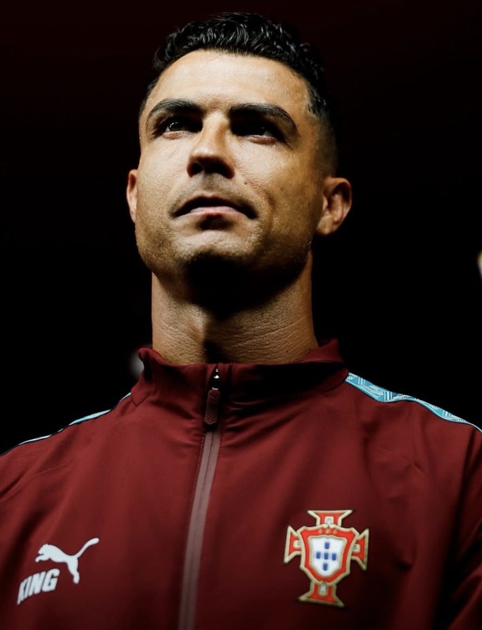 Cristiano Ronaldo in a dark red Portugal tracksuit jacket carrying the national team crest, looking up against a dark background