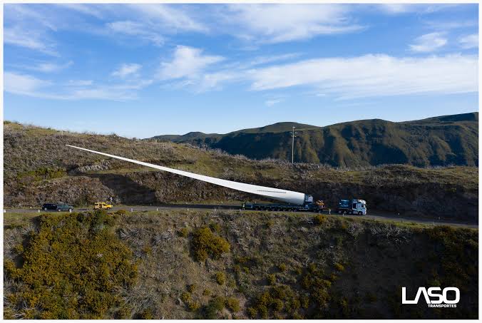 A single wind turbine blade strapped to an extended trailer, photographed side on as it travels along a road