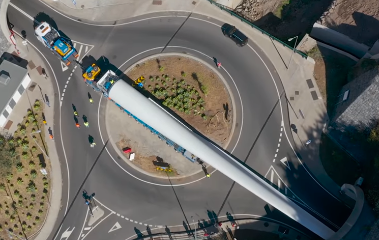 Overhead view of a truck steering a wind turbine blade around a roundabout, the blade overhanging the kerb and the far lane