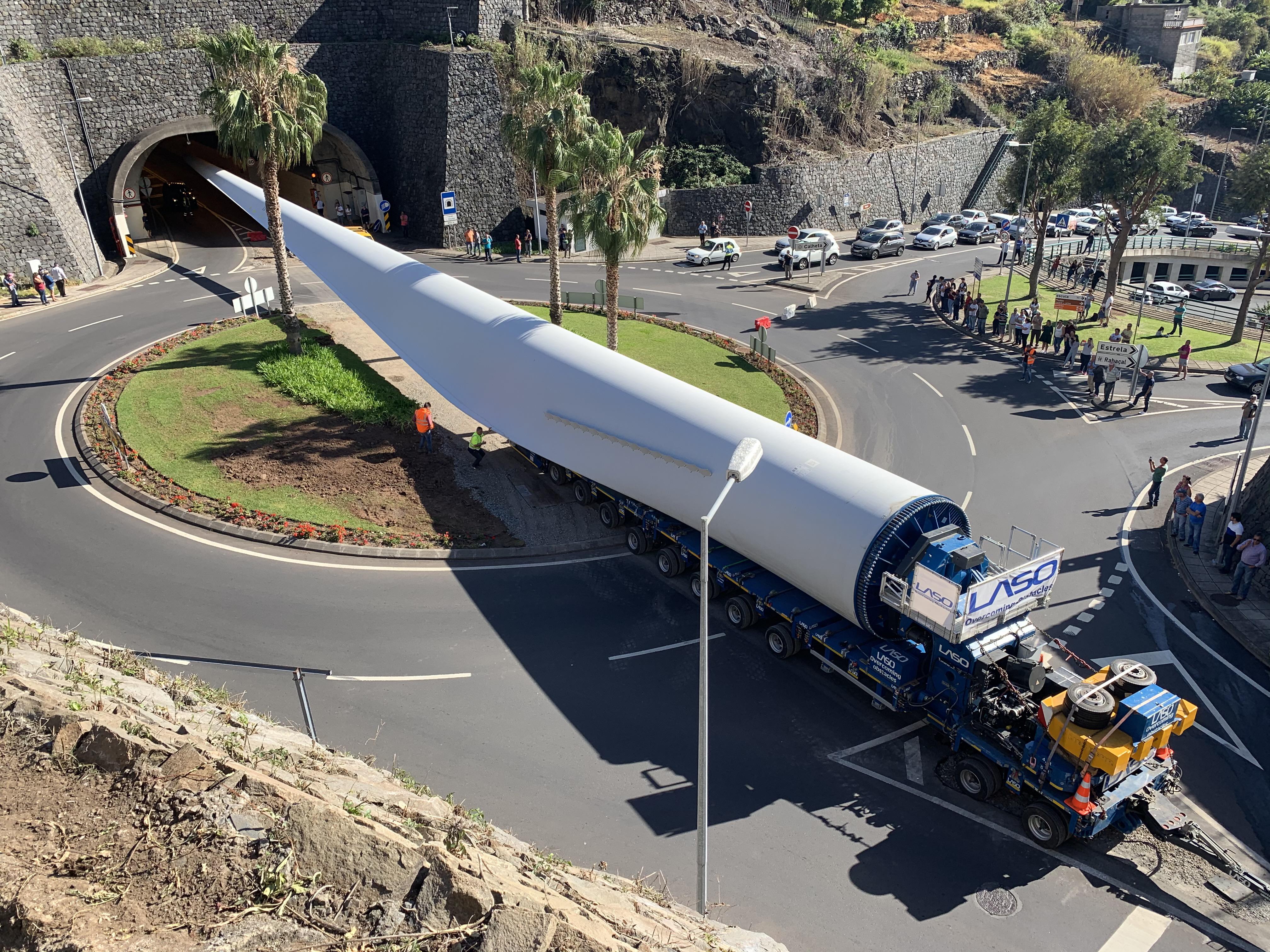 A long low-loader carrying a wind turbine component swinging wide across a roundabout on a Madeiran road near a tunnel mouth